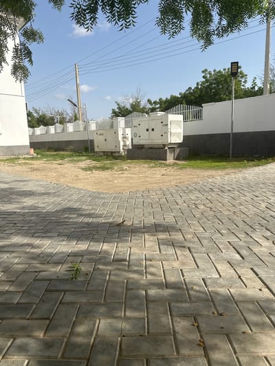 The image depicts an outdoor scene with paved walkways and generators. In the background, a white wall with a decorative fence runs horizontally. Several large, white generators are situated behind the wall, with visible branding. A lamppost with a solar panel is positioned to the right of the generators. A utility pole with overhead wires is also visible. The foreground features a herringbone-patterned paved walkway, with some dry leaves and a small green plant. The sky is blue with scattered clouds, and trees are visible in the distance. This scene is located in the vicinity of Muhammadu Dikko Stadium, Nigeria.