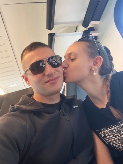 A selfie shows a man and a woman indoors. The woman is kissing the man's forehead. The man is wearing sunglasses and a grey hooded sweatshirt. The woman is wearing a black shirt with a pattern and has braided hair. They are likely on a train or bus, given the interior details. This photo was likely taken in Beckingen, Germany.