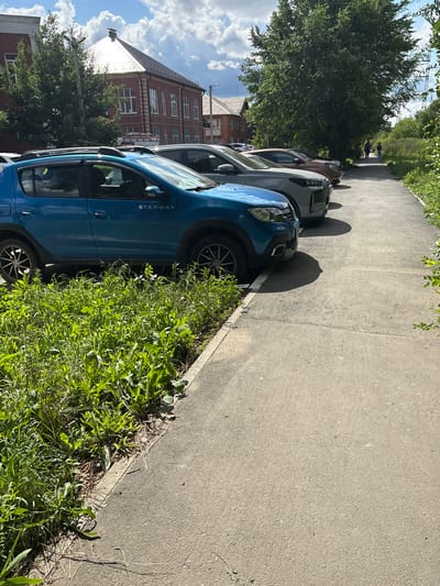 The image depicts a street scene in Orsk, Russia, on a sunny day. In the foreground, lush green grass and weeds grow along the edge of a paved walkway. Several cars are parked in a row on the other side of the walkway, including a blue Dacia Sandero Stepway. Behind the cars, a brick building with white-trimmed windows and a peaked roof stands under a partly cloudy sky. Further down the street, a large tree provides shade, and two individuals, a woman and a child, are walking away from the camera. Power lines are visible in the background.
