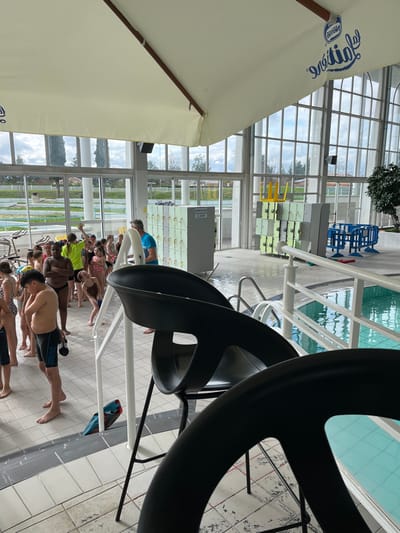 This image depicts an indoor swimming pool facility, specifically the Piscine Antinéa in Andrézieux-Bouthéon, France, identified by the "Nestlé AntínéA" logo on an overhead cream-colored canopy. Approximately 20-25 children, primarily of elementary school age and dressed in swimwear, are gathered on the white and grey tiled pool deck, forming a line. An adult male in a blue shirt is positioned among them, appearing to supervise. In the immediate foreground, two black, high-backed bar stools partially obstruct the view. The background features large, arched floor-to-ceiling windows looking out onto an exterior landscape with trees, green space, and an overcast sky. Inside, along the windowed wall, are several stacks of light green lockers and a grey locker unit displaying a sign that reads "INFORMATION A VENIR". An inverted yellow chair rests on top of one locker stack. To the right, a blue plastic barrier is visible near the pool's edge. The pool itself contains clear, light blue water, bordered by a white railing. A blue kickboard is visible near a floor drain on the deck.