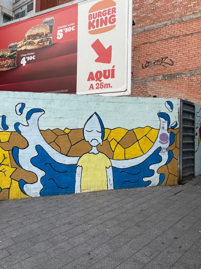 A Burger King advertisement is displayed on a white billboard mounted on a building in Santa Coloma de Gramenet, Spain. The billboard features images of burgers and Coca-Cola, with prices in Euros. Below the billboard, a mural is painted on a brick wall. The mural depicts a sad-looking character with blue and yellow features, surrounded by blue and brown abstract shapes that appear to represent water or a fluid. To the right of the mural, a metal door is visible, and further right, a brick wall with graffiti is present. The ground in front of the mural is paved with grey cobblestones.