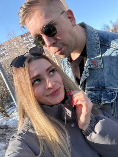 A daytime outdoor photograph captures two individuals, an adult female and an adult male, in a close-up, slightly upward-angled composition. The female, positioned in the foreground, presents with long, straight blonde hair, blue-green eyes, and wears a grey puffer jacket. Dark sunglasses rest on top of her head. Her left hand is raised near her neck, displaying long, pointed red fingernails. She looks upwards towards the male with a slight smile. The male, positioned behind and to the right of the female, has short light-colored hair and wears dark, round sunglasses. He is dressed in a denim jacket over a dark t-shirt, with a small red pin visible on his left lapel. He looks downwards towards the female with a neutral expression. The background reveals an urban environment featuring patches of snow on the ground, bare trees, and a multi-story residential building. A light pole and a fence are also partially visible on the left. The scene is situated in the city of Reutov, Russia.