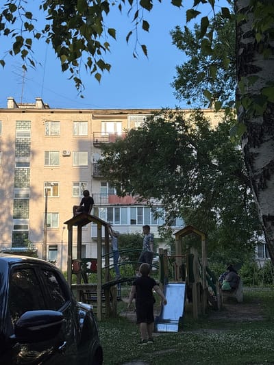 This image captures a playground scene in front of a multi-story apartment building, likely in Sarov, Russia. The building appears to be constructed of light-colored brick and has numerous windows, some with balconies. A large tree with green leaves partially obscures the view, and its branches frame the top of the image.
Several people are visible on and around a wooden and metal playground structure. One person in a black shirt and shorts is sitting on a section of the playground equipment, while another, wearing a light-colored shirt and dark pants, is in a more dynamic pose, seemingly reaching out. A third individual in a patterned shirt and dark shorts is also present near the playground. The lighting suggests it is daytime, with sunlight illuminating the building and parts of the playground. The sky is clear and blue. The overall atmosphere appears relaxed, with the individuals engaged in activities on the playground. A dark car is parked in the foreground, its reflection visible in its side mirror.