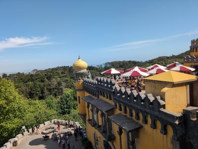 The image depicts a view from a high vantage point at Pena Palace, located in Sintra, Portugal. The foreground features the yellow exterior of the palace, including the battlements with a crenelated design. Red and white umbrellas are visible on the palace rooftop, indicating a dining or relaxation area where people are seated. To the left of the frame, a golden dome rises above a tower. In the courtyard below, a gathering of people is visible, some standing and others seated. The backdrop is filled with a lush green forest stretching towards the horizon. The sky is clear blue with some wispy white clouds.