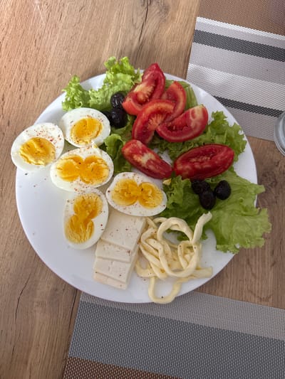 A top-down, eye-level, medium shot captures a white, circular plate of breakfast food resting on a light brown wooden table. The plate contains several halved hard-boiled eggs, some sprinkled with red pepper flakes. A cluster of sliced tomatoes, a bed of lettuce leaves, and a few black olives are also present. To the lower right of the plate, there are slices of white cheese and a tangled mound of string cheese. A placemat with horizontal stripes of brown, gray, and white is partially visible to the right of the plate. The overall scene depicts a casual breakfast setting, likely indoors, with no people visible. The image does not contain any recognizable landmarks or elements that would allow for a definitive determination of its location in Maykop, Russia.