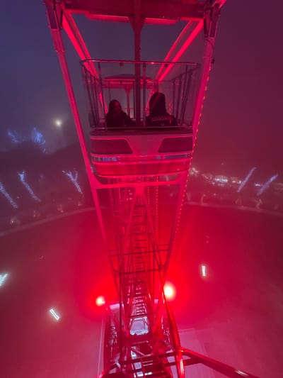 A person sits inside a Ferris wheel car, with shoes prominently displayed. The illuminated red structure of the Ferris wheel dominates the view. Outside, the city is shrouded in fog with faint lights visible. The overall scene is bathed in a strong red light.