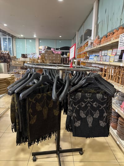 The image depicts the interior of a shop in Acıpayam, Turkey, specializing in handicrafts and home goods. In the foreground, a metal clothing rack holds several dark-colored scarves or shawls with intricate woven patterns, primarily floral designs in lighter shades. These items are hung on black plastic hangers. The floor is tiled in a light beige color.

In the mid-ground, shelves are visible displaying a variety of products. The left side shows boxes and what appear to be textile items. The right side features shelves stocked with terracotta pottery, including vases and bowls, along with other decorative objects and smaller packaged goods. A red sign with white lettering is also attached to a shelf support.

The background reveals more of the shop's interior, including large windows or glass partitions, fluorescent lighting on the ceiling, and additional shelving units filled with various merchandise. The overall impression is of a well-stocked retail environment offering a range of artisanal and decorative items. No people are visible in the image.