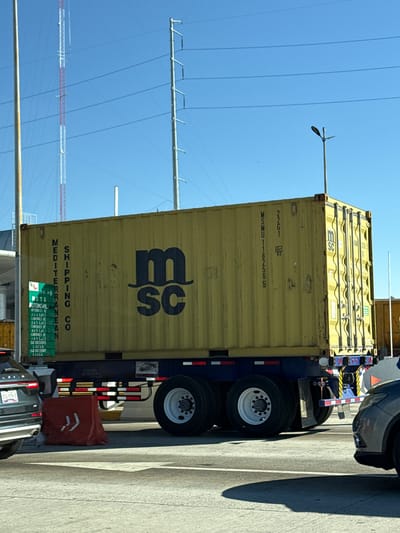 This image captures an outdoor scene on a paved road in Santa María Moyotzingo, Mexico, under a clear blue sky with bright sunlight. The central subject is a large, yellow corrugated shipping container, prominently displaying the black "m sc" logo of MSC (Mediterranean Shipping Company) on its side. Vertical text reads "MEDITERRANEAN SHIPPING CO." Another visible side of the container is marked "MSMU 1182565" and "22G1," along with a "tlc 83" symbol. The container rests on a blue tandem-axle trailer, which features white multi-spoke wheels and red and white reflective safety striping along its lower edge.
In the foreground to the left, the rear section of a dark-colored SUV is visible, displaying a Mexican license plate partially reading "XTC-579-D." Adjacent to this vehicle and the trailer is a red plastic road barrier marked with white chevron symbols. To the right, the front portion of another dark-colored vehicle is partially visible.
The background features a tall, red and white striped lattice communications tower on the left, with power lines or communication cables spanning the sky. Another tall, thin pole, possibly a utility pole, is also visible. On the right, a utility pole with a street light fixture is present. A green rectangular sign, partially obscured, is attached to a pole near the front of the container, displaying text in Spanish that appears to categorize vehicles (e.g., "AUTOMOVIL," "CAMIONES," "CARGA"). Strong shadows are cast on the light-colored paved road surface, indicating the sun's position. No individuals are visible in the scene.