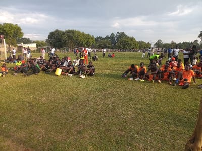 This image depicts an outdoor scene on a grassy field in Chevaywa ward, Kenya, under a predominantly overcast sky. In the foreground, two distinct groups of individuals are seated directly on the ground. The group positioned to the left consists of approximately 25 individuals, primarily young males, many wearing black and white sports uniforms; some are in varied casual attire. A single yellow plastic bucket rests adjacent to this group. The group on the right comprises approximately 25 individuals, uniformly clad in orange sports uniforms. Many individuals in both groups wear athletic footwear. In the mid-distance and background, numerous individuals, including adults and children, stand along the field's periphery. Visible structures in the far left background include a white goalpost and a cluster of single-story buildings with corrugated roofs. A dense line of trees forms the horizon in the distant background. No active dynamic movement is occurring among the seated or standing subjects.
