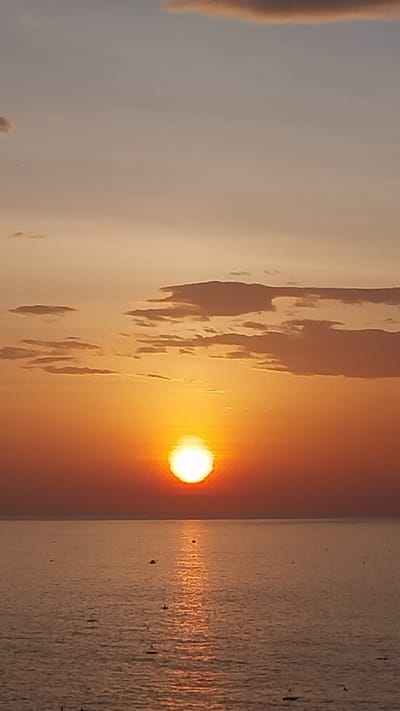 A vertical, medium shot captures a tranquil sunset over a calm body of water. The sun, a bright orb, is positioned in the center of the frame, casting a golden glow that saturates the sky with hues of orange and peach. Wispy clouds, tinged with the same warm colors, are scattered across the upper portion of the image. The water below reflects the vibrant sky, creating a shimmering pathway of light extending from the sun to the foreground. Several small boats are visible on the water, their dark silhouettes contrasting with the bright reflection. The horizon line is sharp and distinct, separating the sky from the sea. The overall atmosphere is serene and peaceful, indicative of a quiet evening in a coastal area, potentially in Tuy Phong 4, Vietnam, known for its beautiful sunsets.