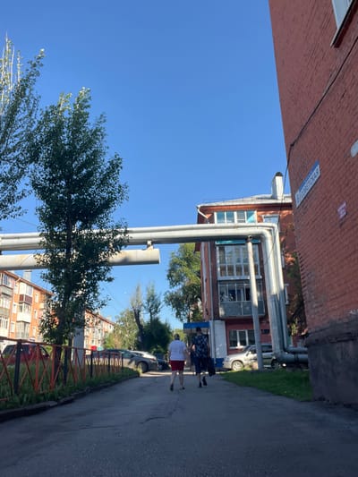 The image depicts a street scene in the city of Kiselyovsk, Russia, under a clear blue sky. A red brick building, part of a larger apartment complex, is on the right side of the frame, with a street sign labeled "6 СОВЕТСКАЯ" (6 SOVIETSKAYA) attached to its wall. To the left, another apartment building with multi-colored balconies is visible. A network of large, grey pipes runs horizontally across the middle of the image, supported by brackets on the red brick building. In the foreground, two individuals are walking away from the camera down an asphalt path. One person is wearing a white t-shirt and red shorts, and the other is wearing a patterned shirt and dark shorts, carrying a black bag. Several cars are parked on the left side of the path, behind a low, orange and black metal fence. Trees with green foliage are present on both sides of the scene.