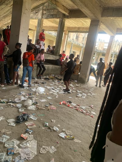 The image depicts approximately 25-30 individuals, predominantly young adults, congregated within the ground level or an open-air floor of a large, unfinished or derelict multi-story concrete structure in Uyo, Nigeria. The environment is characterized by exposed concrete pillars and beams supporting a raw concrete ceiling. The dirt and concrete floor is heavily littered with various items, including numerous plastic bags, food wrappers, paper fragments, and a visible strip of red and white striped barrier tape.
Individuals are observed standing, walking, and sitting on a low concrete ledge or elevated platform. Their attire is casual, comprising t-shirts, jeans, shorts, and footwear such as sandals or sneakers; several individuals are carrying backpacks or shoulder bags. Bright natural light enters the space through large openings between structural elements, revealing further unfinished or dilapidated concrete buildings in the background. No distinct organized activity or interaction is evident among the individuals; the scene suggests a casual gathering or waiting area. The lower right foreground is partially obscured by the dark dreadlocks and shoulder of an unseen individual.