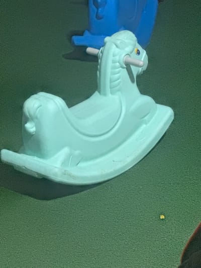 A light teal rocking horse toy sits on a green surface. The rocking horse is positioned on its rockers, facing right, with its mane and tail sculpted. The toy has yellow and blue eyes, and light pink handles protruding from its head. In the background, out of focus, is a portion of a larger blue plastic toy. The green surface appears to be a textured playground matting or artificial turf. A small, yellow debris item is visible on the green surface to the right of the rocking horse. No people are visible in the image. The location is not discernible from the image itself.
