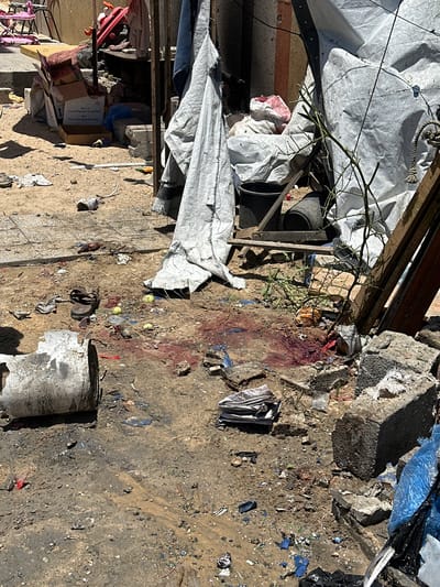 In the city of Gaza, Palestinian Territories, this image depicts a scene of apparent destruction and disarray. The ground is a mixture of dirt and debris, with scattered remnants of what appear to be building materials, such as concrete blocks and twisted metal. Several items are visible on the ground, including a single sandal, pieces of fabric, and what seem to be discarded food items or fruit, some of which are stained red, possibly with blood. There are also plastic bags and crumpled paper. In the background, there are structures made of corrugated metal and fabric sheeting, suggesting makeshift shelters or storage areas. The overall impression is one of a severely impacted area, possibly following a conflict or disaster.