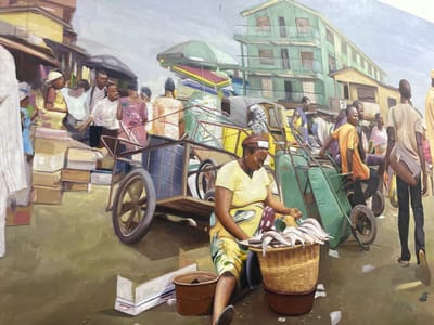 The image depicts a bustling outdoor market scene in Alaguntan, Nigeria. In the foreground, a woman, wearing a yellow top and a patterned skirt, is seated on the ground. She is actively arranging multiple silver fish, resembling catfish, within a large woven basket that rests on a dark basin. To her left, a brown bucket and a discarded white cardboard box lie on the unpaved ground.
The midground is populated with numerous individuals and market transportation. A man in an orange shirt pushes a green, single-wheeled wheelbarrow towards the right side of the frame. To his right, another man, dressed in dark attire, walks away from the viewer, carrying a light-colored shoulder bag. Near the center-left, a large, two-wheeled wooden cart, loaded with items including a prominent blue drum and various bundled goods, is being maneuvered. Several other market-goers and vendors are visible, walking, standing, and interacting. Piles of cardboard boxes are stacked along the left side, indicating market stalls or storage areas. The ground surface is uneven and unpaved, with visible dirt and scattered debris.
The background features urban architecture characteristic of a market area. On the far left, lower structures or market stalls with brown roofs and awnings are visible. Prominently in the center-right background stands a multi-story building with a light green facade and white trim, featuring multiple windows and balconies. Adjacent to it on the right, a shorter, beige-colored building is present. The sky above is a muted, light gray.
