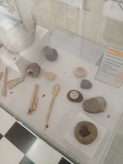 The image shows an exhibit titled "Prácticas Inhala" (Inhale Practices) from an unidentified museum, presumably in Villa Carlos Paz, Argentina. The display features six numbered items related to inhaling practices. Number 1 shows what appears to be a "Mortero Antropomo" (Anthropomorphic Mortar), while number 2 exhibits "Morteros y Manos" (Mortars and Hands). "Molinillo" (Grinding Mill) is designated as number 3, and "Espátulas Óseas" (Bone Spatulas) are numbered 4. Number 5 shows "Tubos" (Tubes), and "Espátula Anatrópica" (Anatropic Spatula) is number 6. All objects are exhibited in a glass case, on a white background. The case sits on a floor with a black and white checkered pattern.