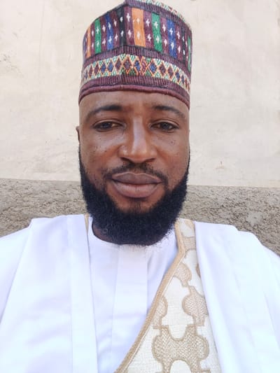 The image, taken in the city of Doma, Nigeria, depicts a headshot and upper torso of an adult male. He has dark skin, a full black beard, and dark eyes, looking directly into the camera with a neutral expression. He wears an embroidered, multi-colored cap featuring vertical stripes in blue, green, orange, and white patterns over a dark maroon base, along with a horizontal band of geometric designs. He is dressed in a white, traditional-style garment with a simple collar. A separate piece of cream-colored fabric, intricately patterned with repeating tan geometric shapes, is draped over his right shoulder. The background consists of a plain, light beige wall with a subtle texture.