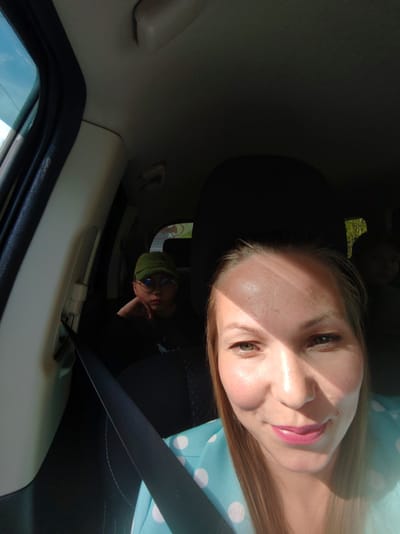 A woman in a blue and white polka dot shirt takes a selfie inside a car. She is wearing a seatbelt. The back seat holds a person wearing a green cap and glasses, looking at the camera. Another person is partially visible behind the front passenger headrest. The interior of the car is visible, including the window frame and roof. Based on the tree visible through the back window, the car may be traveling in a rural or suburban environment near the city of Vyl'gort, Russia.