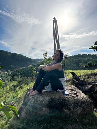 A woman sits on a large rock outdoors, embracing her knees. She wears a dark tank top and dark leggings, with sunglasses perched on her head. The sun shines brightly in the sky, which is filled with white, wispy clouds. In the background, a lush green mountainside dotted with trees rises, suggesting a rural or natural setting. A structure made of bamboo poles stands vertically near the woman, its purpose unclear. The lighting and shadows indicate it is daytime. The woman's posture and relaxed expression convey a sense of peace and contemplation. The scene appears to be in Thôn 3, Vietnam, given the mountainous terrain and vegetation.