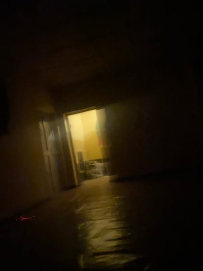 The image shows a dimly lit hallway with a door open at the end, revealing a yellow-lit room. The hallway walls are textured and appear to be painted. The floor is covered with a reflective material, possibly plastic sheeting, which catches the light. In the foreground, to the left, a small, brightly lit red object is visible on the floor. The overall atmosphere is dark and somewhat eerie, with limited visibility. The specific location in Barikin Jirga Summa, Nigeria cannot be determined from the visual information provided in the image.