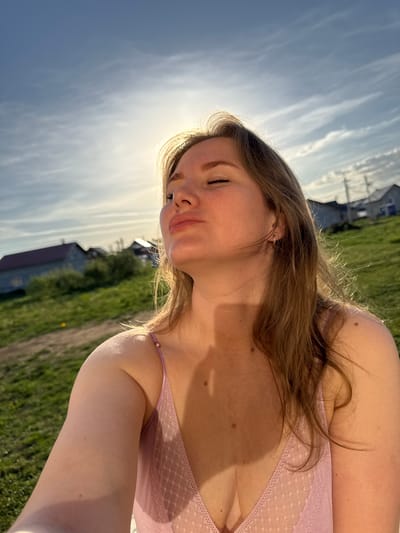 The image depicts a woman outdoors. She has fair skin and light brown hair. She is wearing a pink top with lace trim. The background shows a grassy field and buildings, consistent with a rural or suburban setting. The sky is visible in the background with a few clouds and the sun positioned above the woman's head, which is in the city of Ферма, Russia.