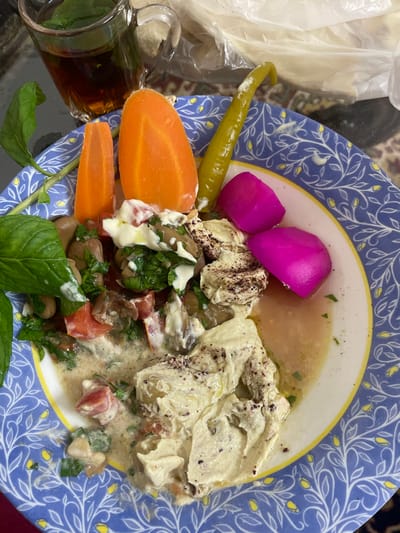 The image displays a close-up, top-down view of a meal presented on a blue and white patterned ceramic plate with a yellow rim, situated within an indoor setting in the city of Al-Qanawat Municipality, Syria. The plate contains Foul Medames (fava beans) mixed with chopped tomatoes, green herbs, and a creamy white sauce, with visible olive oil. Adjacent to this, a portion of light-colored mashed chickpeas or hummus is topped with sumac and olive oil. Accompanying items on the plate include two distinct, oblong slices of cooked orange carrot, one long green pickled pepper, and two bright pink pickled turnip pieces. Fresh green mint sprigs serve as a garnish on the left side of the plate. To the upper left, a clear glass cup with a handle contains a dark amber liquid, consistent with tea, featuring green mint leaves floating on its surface. A crumpled transparent plastic bag is partially visible in the background, behind the plate. The entire arrangement rests on a subtly patterned surface, possibly a textile or mat.