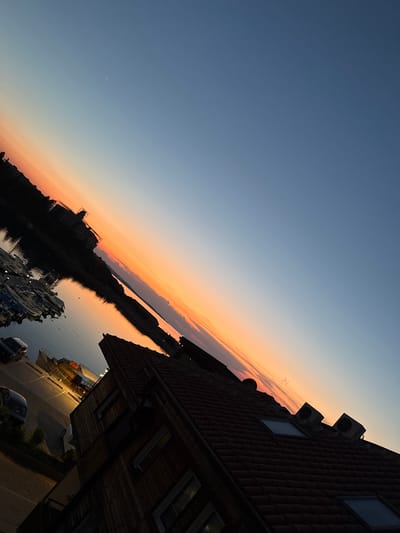 The image depicts a sunset scene in Sozopol, Bulgaria. The sky is a gradient of deep blue at the top transitioning to warm orange and pink hues near the horizon, indicating dusk. The sun is not directly visible, but its light creates a dramatic reflection on the calm water of a harbor.
In the foreground, to the right, is the side of a building with a red tiled roof. Several windows are visible, and two air conditioning units are mounted on the wall. The building appears to be illuminated by artificial lights, suggesting it is nighttime or that the interior lights are on. Below the building, a paved area with parked cars and a street are visible.
Across the water, a silhouetted landscape stretches along the horizon. A prominent structure, possibly a building or monument, is outlined against the colorful sky. A cluster of boats are docked in the harbor, their shapes also silhouetted. The overall mood is serene and picturesque, capturing the quiet beauty of a coastal town at sunset. No visible text is present in the image.