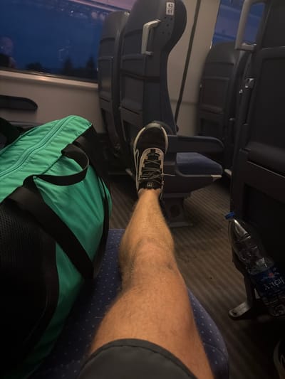 The image depicts the interior of a train carriage. A person's leg, clad in a black and white sneaker, is extended forward, resting on the seat in front. To the left of the leg is a large, teal-colored duffel bag with black straps. The train seats are upholstered in dark blue fabric. Through the window on the left, the dark sky and silhouetted trees are visible, suggesting it is either evening or early morning. A plastic bottle of water with a blue cap sits on the floor to the right. The flooring is a dark, ribbed material. There are no identifiable landmarks or context to place the scene specifically in Gavrailovo, Bulgaria.