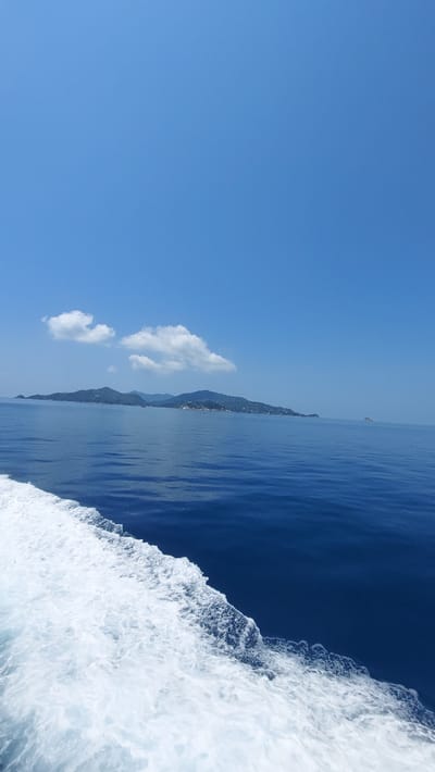 The image shows a body of water, most likely the ocean, with white water churning at the edge of the frame, likely from a boat moving through the water. In the distance, there is a coastline with mountains visible. A few white clouds are visible in the clear, light blue sky. The photo appears to be taken during the daytime. There are no people immediately visible. The location in Ko Tao, Thailand cannot be definitively determined from the given image.
