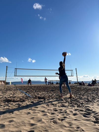 The image depicts a sunny day at a sandy beach, likely in Tolyatti, Russia, given the context. The sky is a clear, vibrant blue with a few scattered white clouds. A volleyball net is set up on the sand, indicating a beach volleyball game is in progress or has recently occurred.
The main subject is a person, silhouetted against the bright sky, mid-air as they dive to hit a volleyball. They are wearing a dark long-sleeved shirt and light-colored leggings. Their body is extended, one arm reaching out to make contact with the ball, which is also visible.
In the background, other people are present on the beach, some playing or spectating near the net, while others are further back, possibly relaxing or walking along the shoreline. The sea or a large body of water is visible in the distance, with a hazy horizon. The sand is textured with ripples and shadows, suggesting the sun is high in the sky, likely late morning or early afternoon.
There is a red flag planted in the sand with some white text on it, but it is partially obscured and illegible. Another smaller white flag is visible further down the beach. The overall mood is active and energetic, capturing a moment of athletic play on a beautiful, clear day.