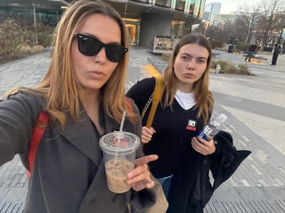 Two young women pose in front of the Denver Art Museum. One holds a "smartwater" bottle and wears a black sweater with an "ART" logo, while the other holds an iced coffee in a plastic cup and wears sunglasses and a gray coat. They stand on a paved area with trees and other buildings in the background. The image is rotated and slightly blurry.