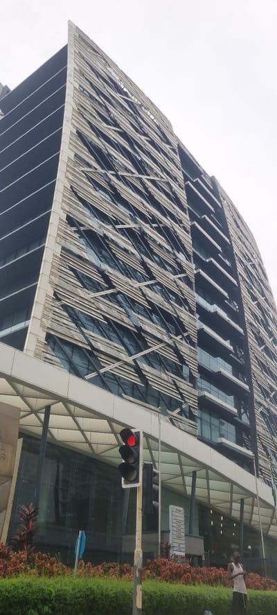 The image shows a low-angle view of a modern, tall office building in Ikoyi, Nigeria. The building has a distinctive facade with horizontal white fins that create a textured, angular pattern over dark glass windows. The top of the building is sharply angled and protrudes outwards. In the foreground, a traffic light with a red light illuminated is visible, along with a street sign. A person wearing a white tank top and dark shorts stands to the right of the traffic light. Lush green bushes and red-leafed plants line the area in front of the building. The sky is overcast, suggesting a cloudy day. The overall scene depicts an urban environment with a focus on the architecture of the building and its immediate surroundings.