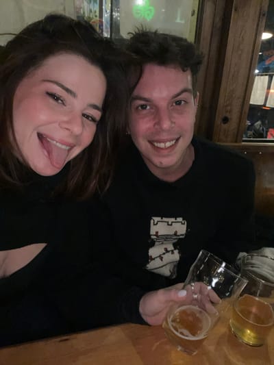 A man and woman pose closely, smiling. The woman sticks out her tongue. Both are wearing black shirts. The man's shirt has a white and red design. Three glasses of beer sit to the right. The scene is indoors with a wooden background, possibly a bar. The lighting is dim and warm.
