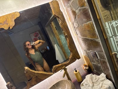 A woman in a green one-piece swimsuit and sunglasses smiles at her reflection in a large, irregularly shaped mirror with a wooden frame. She is taking a selfie with a phone. The bathroom features stone walls, bamboo accents, and a wooden countertop. Visible objects include a faucet, a soap dispenser, a small decorative vase with flowers, and a folded white towel with embroidery. The room appears to be a high-end spa or hotel bathroom, possibly located in Nam Hoa Lư Ward, Vietnam, given the apparent architectural style and bamboo decor.