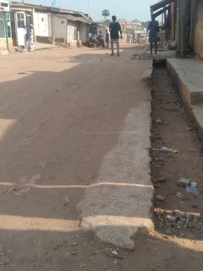 The image displays an unpaved, reddish-brown dirt road in Agege, Nigeria, under clear daylight conditions, with shadows indicating strong sunlight. The road surface is uneven, featuring a lighter, compacted strip running horizontally across its midsection.

On the left side of the road, a series of low-rise buildings line the street. The nearest structure has a white door and a partial blue awning or sign. A person in a patterned blue and white dress stands near this building's entrance. Further down the left, a small group of approximately three to five individuals is gathered near a small structure or stall.

On the right side of the road, a concrete drainage ditch, partially filled with dirt and scattered debris, extends along the length of the visible area. Adjacent to the ditch is a dark-colored building with an exposed wooden overhang or support structure. A person wearing a dark blue shirt and patterned shorts stands near the ditch, facing towards the left.

In the mid-ground, a person wearing a dark shirt and light trousers stands with their back to the camera, positioned slightly to the right of the road's center, facing down the street. In the background, additional buildings, some appearing multi-story, are visible, interspersed with sparse vegetation, including a solitary tree. No explicit interactions or specific actions are discernible beyond individuals standing or moving along the street.