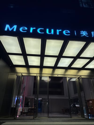 A nighttime, low-angle exterior shot shows the entrance to a Mercure hotel in Huangpu District, China. The hotel's name, "Mercure," is illuminated in bright blue neon letters against a dark background. Above the entrance is a modern canopy with a grid of illuminated panels. The entrance itself features automatic glass doors that are partially open, revealing a glimpse of the hotel's lobby. Inside, elements of the lobby are visible, including decorative lighting fixtures, furniture, and what appears to be a reception area. The overall impression is of a well-lit and contemporary hotel entrance at night.