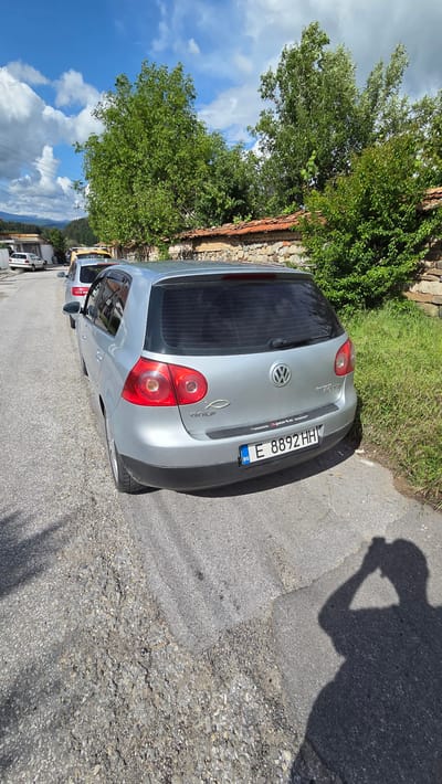 The rotated image captures the rear-right view of a silver-grey Volkswagen Golf (likely a Mk5 model) parked on an asphalt road in Bania, Bulgaria, under clear daylight conditions. The car's tailgate features the circular Volkswagen emblem, "GOLF" badging on the lower left, and "2.0 TDi" on the lower right. A small "sports" decal is located above the license plate, and a silver fish symbol (ichthys) decal is affixed below the "GOLF" lettering.
The vehicle displays a Bulgarian license plate: "E 8892 HH," with the "BG" country identifier on the left. The rear window of the car is tinted. To the left of the vehicle, a stone and brick wall is visible, overgrown with dense green foliage and trees that extend upwards into a bright blue sky dotted with white clouds. In the background, hints of distant mountains and other structures suggest a village or town setting.
The road surface is asphalt, exhibiting signs of wear with cracks and patches of gravel. A prominent shadow of a person, holding what appears to be a camera or phone, is cast on the road in the foreground, indicating the photographer and suggesting a sunny mid-day or afternoon. The scene depicts a serene outdoor environment.