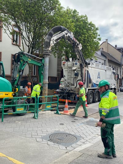In Bischheim, France, a team of construction workers are conducting underground maintenance. A large white vacuum truck with "SOBECA" branding is parked next to the excavation site. One worker in a green and yellow reflective vest, green helmet, and gloves is positioned near the manhole cover. Another worker in a green uniform and red helmet leans over the truck's controls. An excavator and green safety barriers, also bearing the SOBECA logo, surround the open excavation site. A third worker in an orange helmet and vest is visible within the excavation. The surrounding environment includes residential buildings and trees.