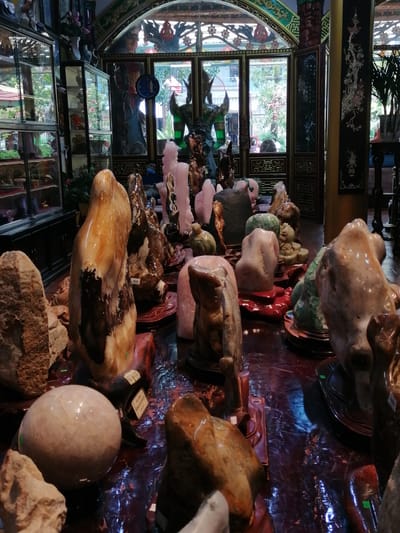 This is a forensic, matter-of-fact description of an indoor scene, likely a shop or display area, featuring numerous polished stones and decorative objects. The environment appears to be well-lit, with sunlight filtering through large arched windows at the rear, revealing an outdoor area with trees. The foreground is dominated by a collection of various stones, some large and abstractly shaped, others smaller and more defined, displayed on dark red wooden stands. These stones exhibit a range of colors including earthy browns, whites, pinks, and greens, with polished surfaces reflecting the light. In the background, glass display cabinets line the walls, filled with more objects, some appearing to be vases and other ornamental items. Two figures are partially visible: one man wearing a blue mask is seated on the right, and another person is seen reflected in a glass cabinet on the left. The overall impression is that of a store specializing in decorative stones and possibly antiques, located in Xuân Thành, Vietnam.