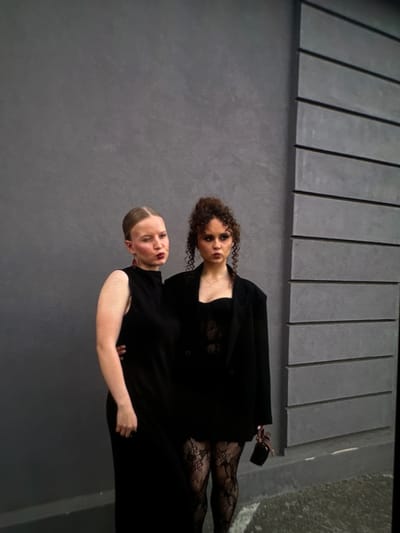 Two young women are standing against a textured gray wall on the left and a ribbed gray structure on the right. The woman on the left has her hair pulled back in a bun, is wearing a sleeveless black dress, and has on large red earrings. She is looking to her left and appears to be winking. The woman on the right has curly brown hair and is wearing a black blazer over a black, lacy top and textured black stockings. She is looking forward with a neutral expression and is holding a small black clutch in her right hand. The background suggests an outdoor urban setting in Novi Sad, Serbia, possibly near a modern building.