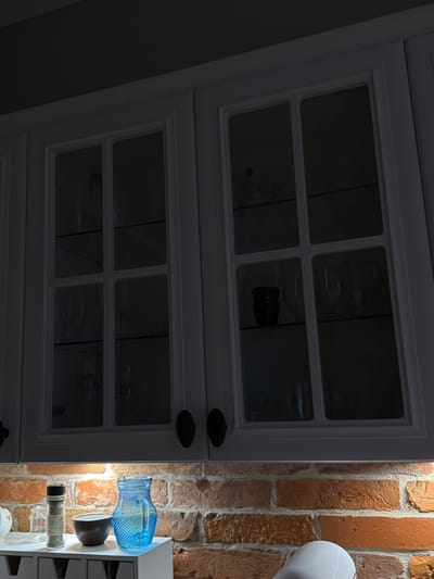 The image shows a close-up of two white cabinet doors with glass panes, set against a backdrop of rustic red brick. The cabinets appear to be mounted on a wall, and the overall scene suggests an indoor setting, possibly a kitchen or dining area.

Inside the upper cabinet, several clear glass objects are visible, though their exact nature is obscured by the lighting and reflections. A dark-colored bottle or container is also present. The lower cabinet has similar glass panes, revealing more clear glass objects within.

To the right of the cabinets, on a white shelf unit attached to the brick wall, are several items. A distinctive bright blue glass bottle with an intricate pattern sits at the top of the visible shelf. Below it is a dark, rounded object, possibly a small bowl or vase. Further down, a clear glass container with a black lid is visible, appearing to hold salt or a similar granular substance, with text that reads "SEA SALT". Below this, other clear glass containers are stacked, hinting at a collection of bottles or jars. The overall lighting suggests it is either evening or a dimly lit interior space, with artificial light illuminating the shelf items. The presence of cabinets and shelves with glassware suggests a domestic or decorative setting. The location context indicates this scene is in Słupsk, Poland.