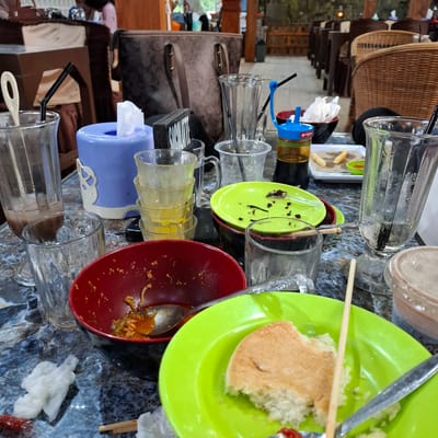 The image shows a messy table at what appears to be a restaurant or cafe in Baroh Musa, Indonesia. The table is laden with used dishes, glasses, and food remnants, suggesting a meal has recently concluded. 

The primary subjects are the objects on the table: several clear glasses, some containing remnants of drinks and straws; a red bowl with leftover food; a bright green plate with a piece of cake and a fork; and a smaller green plate with crumbs. There are also used napkins and a tissue dispenser. Two handbags are also visible on the left side of the table. In the background, wicker baskets and wooden furniture can be seen, hinting at the establishment's decor.

The scene depicts the aftermath of a meal, with the activity implied being dining. The time of day is not explicitly clear, but the lighting suggests it could be daytime. No specific weather conditions are apparent. The presence of Indonesian text on some of the packaging, though not clearly legible, along with the location context, confirms the setting. The overall impression is one of disarray after a gathering or a meal service, possibly indicating a busy period or an upcoming cleanup.