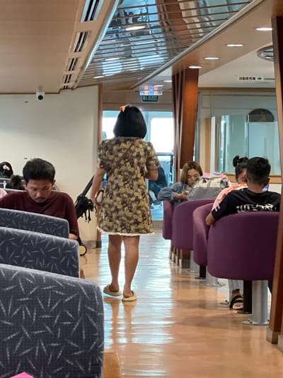 The image depicts the interior of a ferry, likely in the city of Don Sak, Thailand, given its common use as a ferry terminal. In the foreground, blue patterned seats are visible. Several people are seated on purple chairs, facing a walkway. A woman with black hair, wearing a floral dress, walks away from the camera down the walkway. To her left, a man in a maroon shirt is seated, looking down. Further down the walkway, other passengers are visible, including a woman in a denim jacket and another in a black shirt. The ferry interior features overhead lighting and reflective surfaces, suggesting a modern design. Signs in Japanese are visible in the background, indicating the potential international nature of the ferry service.