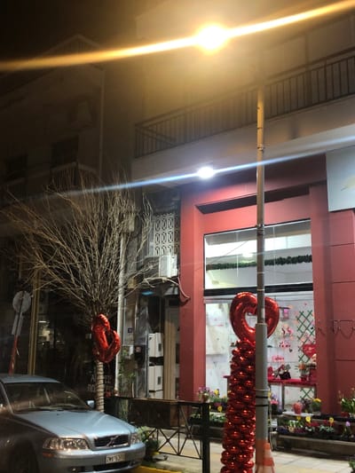 This media file captures a nighttime street scene in Marousi, Greece, featuring buildings, street lighting, decorations, and a parked car. The image is taken at night, evidenced by the darkness and the strong artificial illumination from multiple sources.
In the foreground, a prominent street pole runs vertically through the center, from which hang numerous red, small metallic balloons forming a decorative garland. Attached to this pole, and extending into the windows of a building on the right, are large, red heart-shaped foil balloons, suggesting a festive or romantic occasion, possibly related to Valentine's Day.
To the right, a building with a red-painted facade and large glass display windows is visible. Through these windows, parts of a shop interior can be seen, decorated with a checkered grid pattern and what appear to be flowers or other small items. Below the red facade, there is a section with decorative patterned grilles.
On the left side of the scene, a building with balconies and railings is partially visible, shrouded in the darkness. Two streetlights are prominent: one on the far left emits a warm yellow glow with a strong light flare, while another closer to the center, aligned with the right building, emits a bright white light, also with a significant flare.
Towards the bottom right, a light-colored (possibly silver or light blue) car is parked, with its front end visible. The car's license plate, which reads "ΖΜΛ 5539," is clearly discernible, confirming the Greek location. A bare, leafless tree or large bush occupies a significant portion of the lower and central part of the image, its intricate branches silhouetted against the ambient light. Another red heart-shaped balloon is visible near the bare tree, close to the car. A small, round road sign is also partially visible near the front of the car.
The overall impression is an urban setting at night, adorned for a special occasion. No people are visible, and no specific activity is taking place beyond the stationary scene.