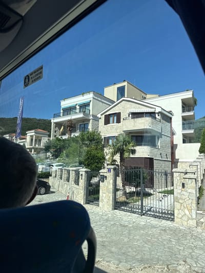 The image shows a view from inside a vehicle, looking out through the windshield at a residential area under a clear blue sky. In the foreground, the back of a person's head and shoulder, wearing a blue garment, are visible, obscuring part of the view.

Outside, a paved driveway leads to a stone fence with an ornate metal gate. Beyond the gate are several modern, multi-story houses constructed with light-colored stone and stucco. One prominent house features multiple balconies with dark railings, stone cladding, and awnings. Palm trees and lush green vegetation are interspersed among the buildings.

A blue flag with white text, possibly advertising a business or event, is visible on a pole to the left of the houses. A yellow sticker with French text, indicating "PORT OBLIGATOIRE DE LA CEINTURE DE SECURITE" (Seatbelt Required), is affixed to the vehicle's window. Several cars are parked behind the fence. The background shows a hillside covered in trees. The scene appears to be in Jošice, Montenegro.