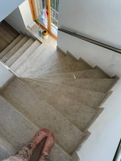 This image shows a person's bare feet with extensive tattoos standing on the bottom steps of a winding staircase. The staircase is constructed of light-colored stone with a marbled texture. A white handrail runs along the right side of the staircase. The walls are white. At the top of the stairs, a window with wooden trim overlooks an outdoor scene with greenery. On the windowsill, there are several objects including a white box, two paper bags, a glass bottle with a green plant, and a beige ceramic vase. The floor next to the top of the stairs appears to be wooden planks. The scene is illuminated by natural light streaming in from the window. The overall impression is that of an interior staircase in a residential or public building in Mtskheta, Georgia.