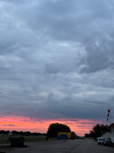 The image depicts a dusk scene with a vibrant sunset over Zgalevo, Bulgaria. The sky is filled with dramatic clouds, with streaks of intense pink and orange light breaking through the dark gray. In the foreground, there is a paved area with several vehicles, including a blue van, a red truck, and a few parked cars. A white building with a red and white windsock on a pole is visible on the right side of the frame. Trees line the horizon, with a dark green industrial-looking tank or silo in the lower left quadrant. The overall atmosphere suggests an airport or industrial area during twilight.