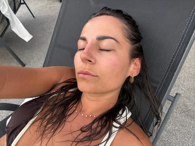 A woman is lying on a grey mesh chaise lounge, outdoors. Her eyes are closed, and her head is slightly tilted back. She has dark, wet hair that is resting on her shoulders. She is wearing a brown and white bikini top and a delicate pearl necklace. A gold hoop earring is visible in her right ear. The background consists of a grey, pebbled surface and a white long-sleeved shirt draped over another chair in the upper left corner. The scene appears to be in Veauchette, France.