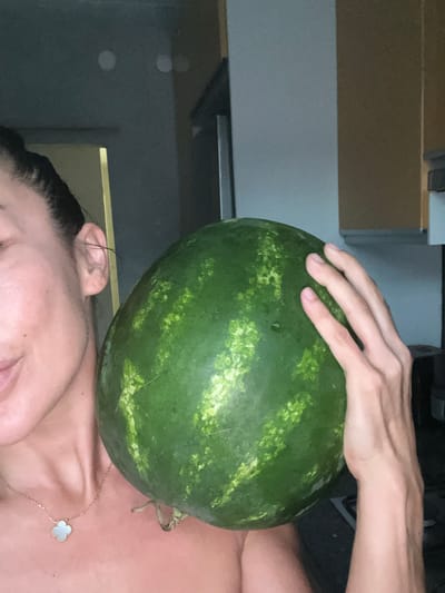 The image shows a close-up of a person's head and shoulder, with a watermelon resting on top of their head. A hand is visible on top of the watermelon, with fingers spread. The person is wearing a necklace with a clover-shaped pendant. The background appears to be indoors, with cabinetry and a wall visible. The lighting suggests it could be daytime. There are no discernible emotions or specific activities beyond the posing with the watermelon. The location context provided is Şişli, Turkey, but no visual cues within the image confirm this.