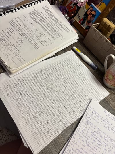 The image shows a close-up of a desk with several notebooks and loose papers covered in handwritten notes, predominantly in Bulgarian. The notes appear to be related to chemistry, with chemical formulas, reactions, and diagrams.
In the foreground, there is a white mug with a floral pattern and a white and blue pen. To the left, a stack of paper with grid lines is filled with dense handwriting. Behind this, a spiral-bound notebook is open, also filled with handwritten notes and chemical equations.
Further back, other notebooks and papers are visible, some with diagrams. In the upper right corner, a wicker basket holds various items, including a framed photograph of two people and a small, brown, star-shaped object. The lighting suggests an indoor setting, possibly a home or study environment. The image does not provide any information or visual cues that would allow for the determination of its location in Bulgarovo, Bulgaria.